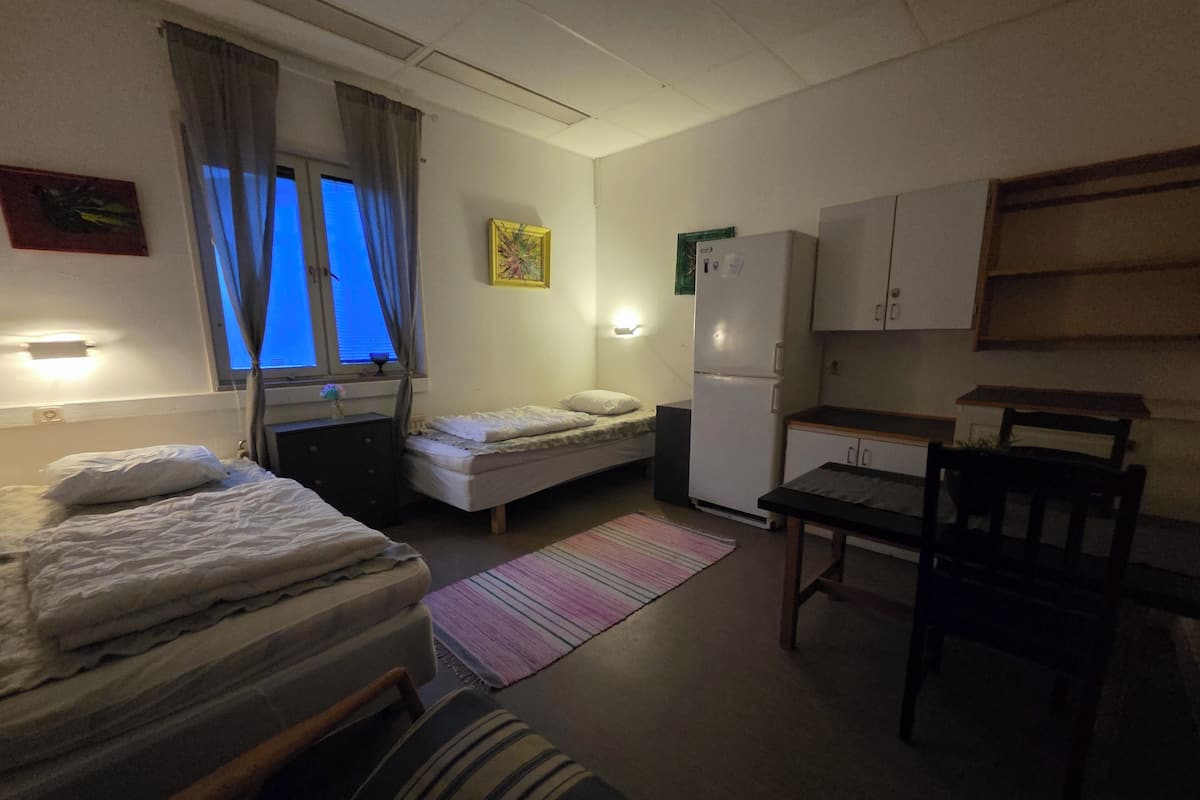 A compact hotel room with two single beds, a small kitchenette, a dining table, and a chair.
