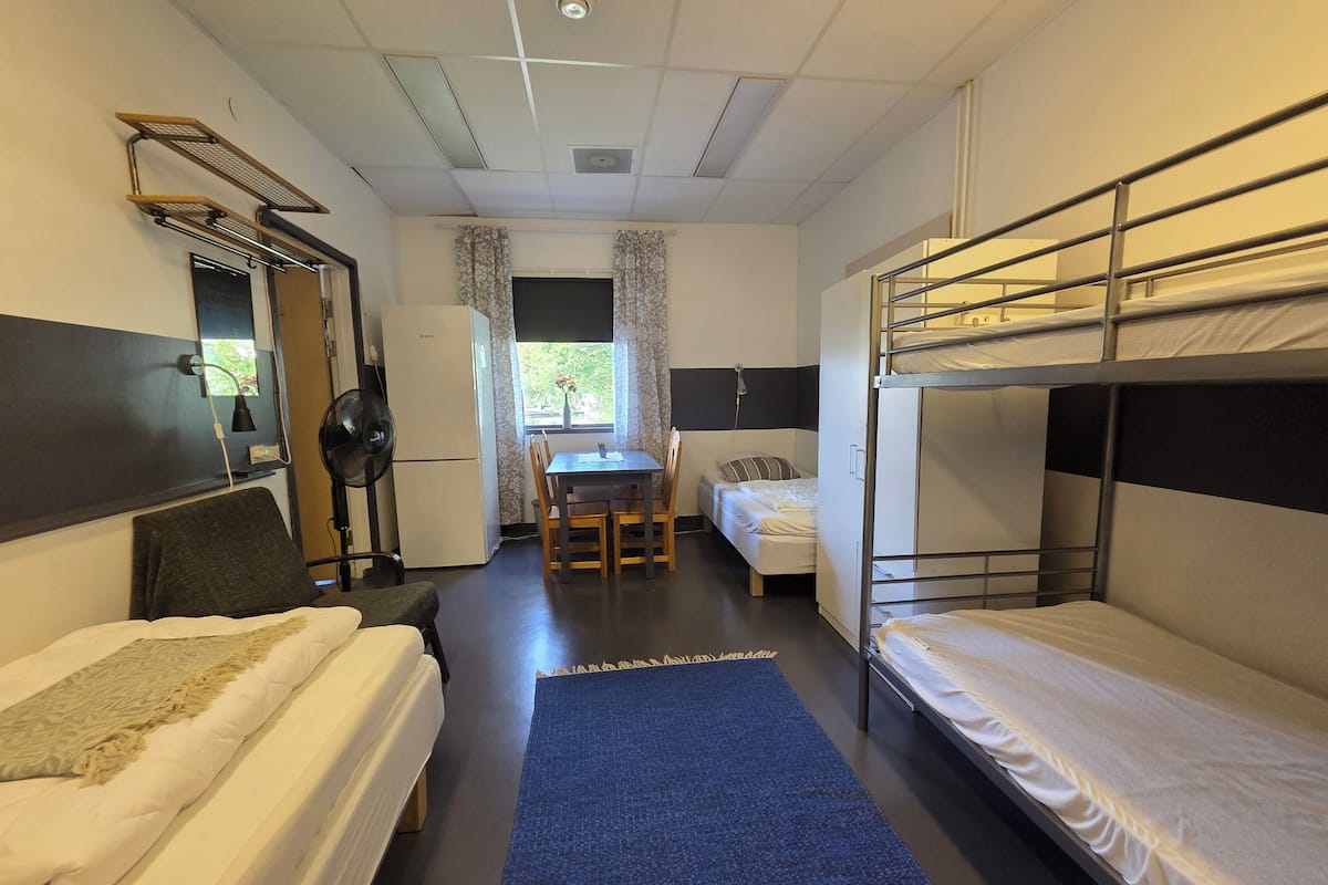 A compact dormitory room with bunk beds, a small kitchenette, a desk, and a window with curtains.