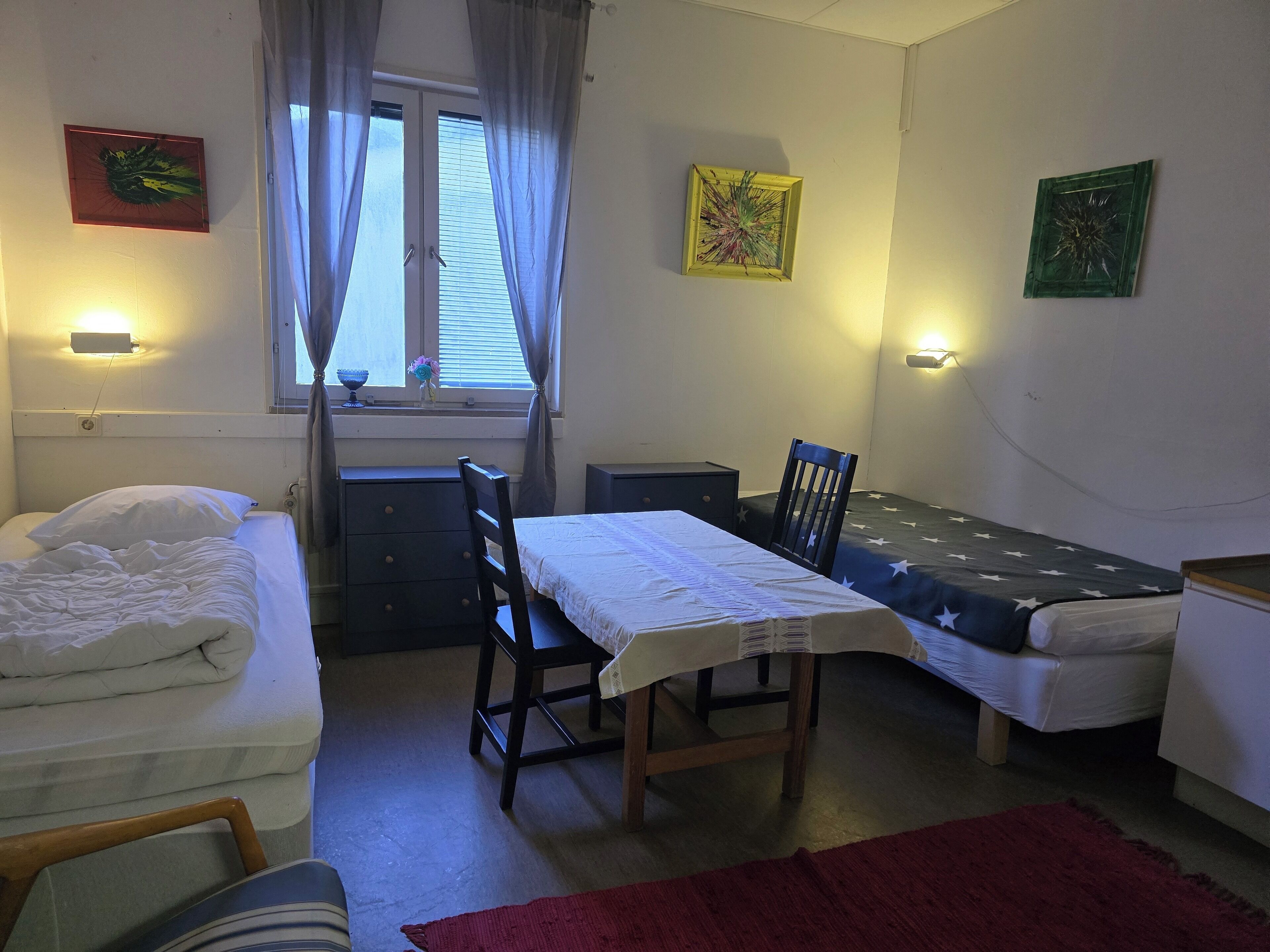 A small, well-lit room with a bed, a desk, a chair, and a window with curtains. There are wall-mounted lights and a small table with a chair.