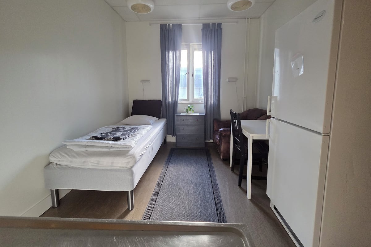 A compact hotel room with a single bed, a small desk, a chair, and a refrigerator.