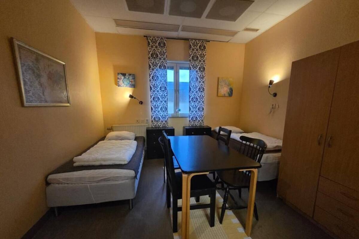 A compact room with a bed, a small table, and a chair. There is a window with curtains, a radiator, and a cabinet.