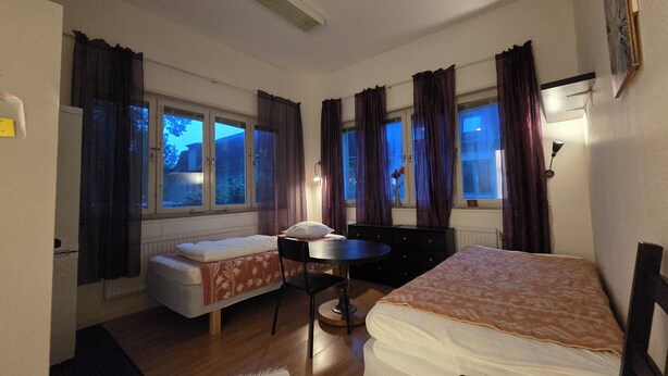 A room with two beds, a small table, and a chair. There are windows with curtains, a radiator, and a wall-mounted lamp. - Svenshögens vandrarhem (Svenshögen)