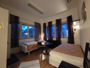 Une chambre avec deux lits, une petite table et une chaise. Il y a des fenêtres avec des rideaux, un radiateur et une lampe fixée au mur.