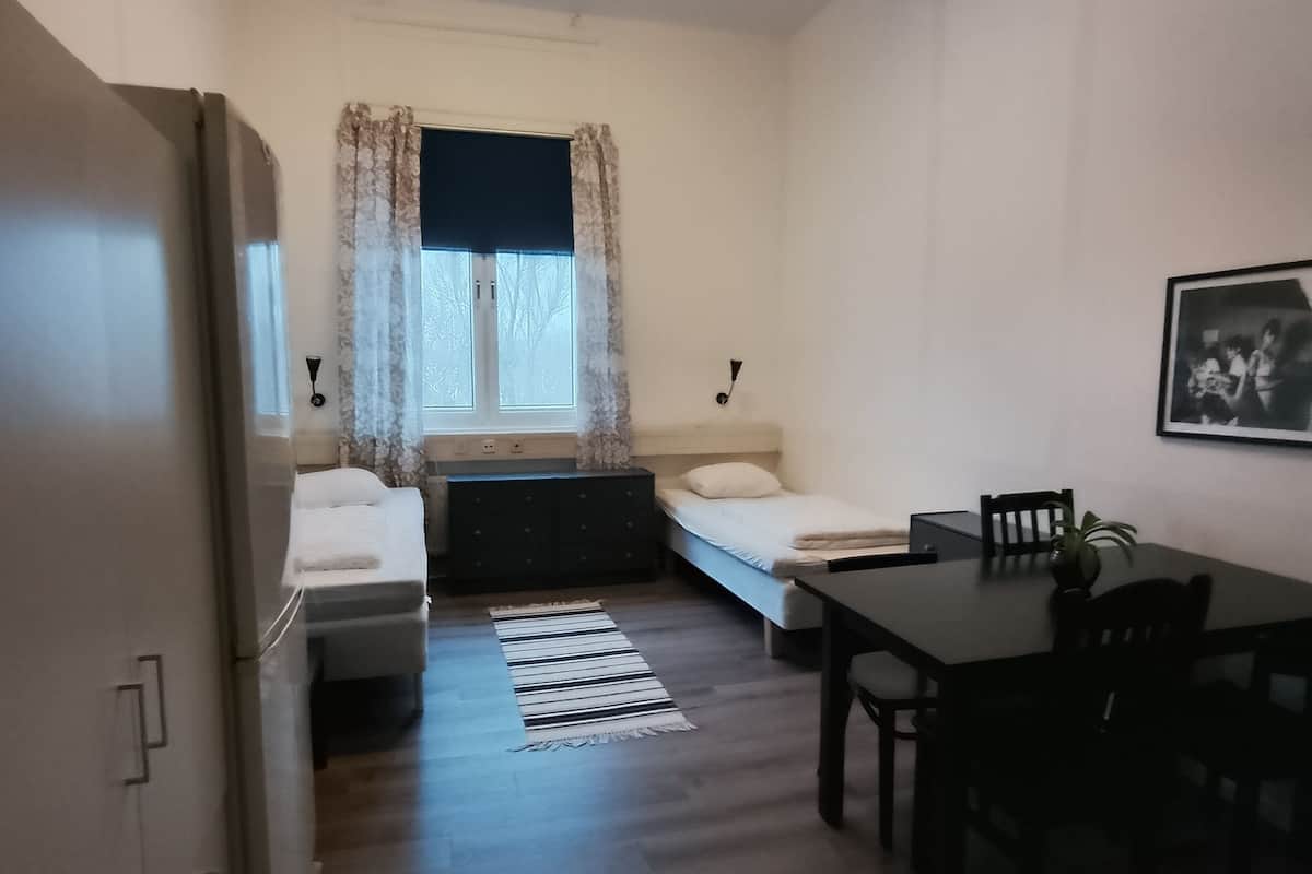 A compact room with a bed, a small table, a chair, a desk, a window with curtains, a radiator, and a wall-mounted picture.