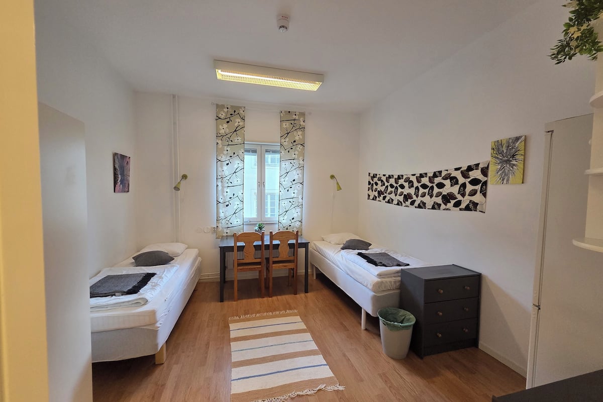 A small, well-lit room with two single beds, a wooden floor, a dining table with chairs, a small trash bin, and a black cabinet.