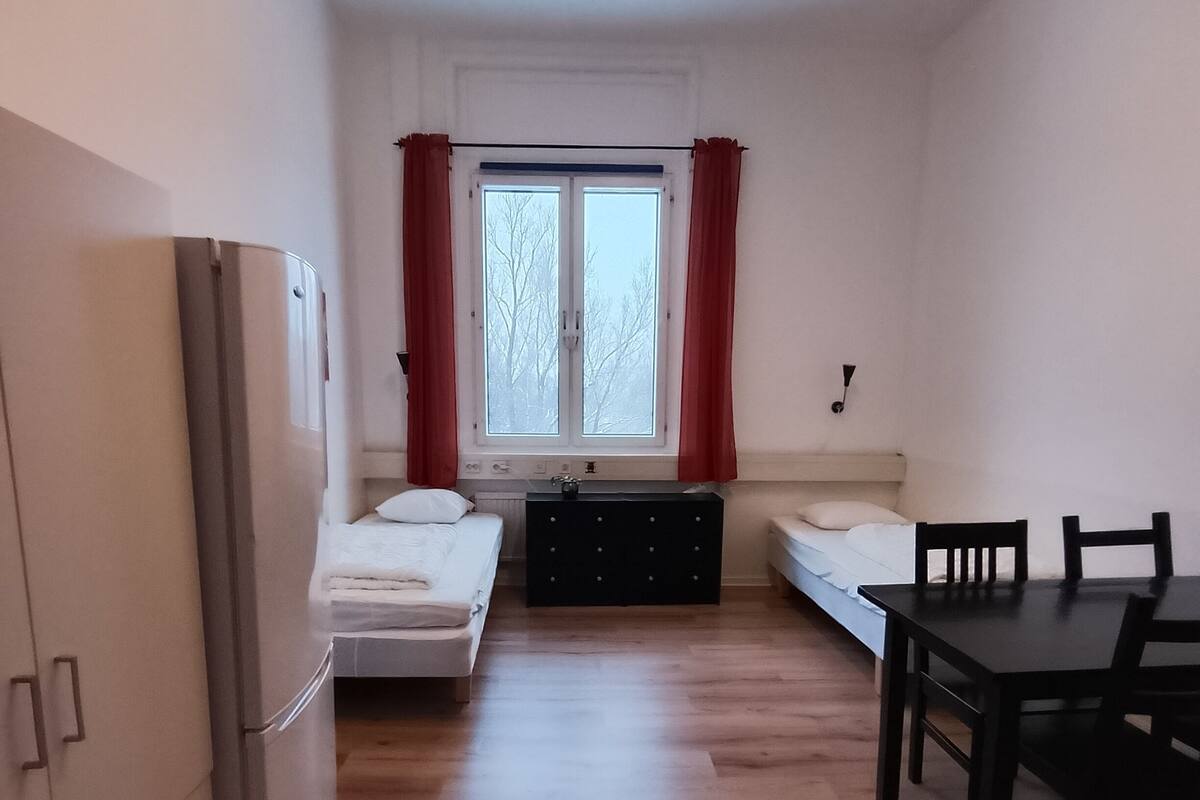 A compact room with a bed, a small table, a chair, a refrigerator, and a window with curtains.