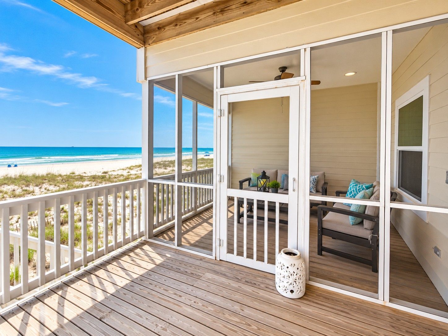 Une terrasse en bois avec vue sur la plage, un coin salon avec des coussins et un vase blanc.