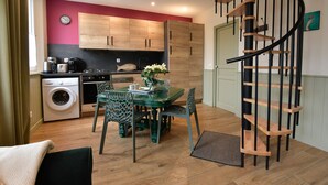 A compact kitchen with wooden cabinets, a washing machine, and a dining area with a green table and chairs. - The Little Cottage in the Bay of Somme (Cahon-Gouy)