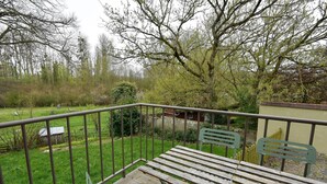 A balcony with a wooden table and chairs, overlooking a grassy area with trees and a small building. - The Little Cottage in the Bay of Somme (Cahon-Gouy)
