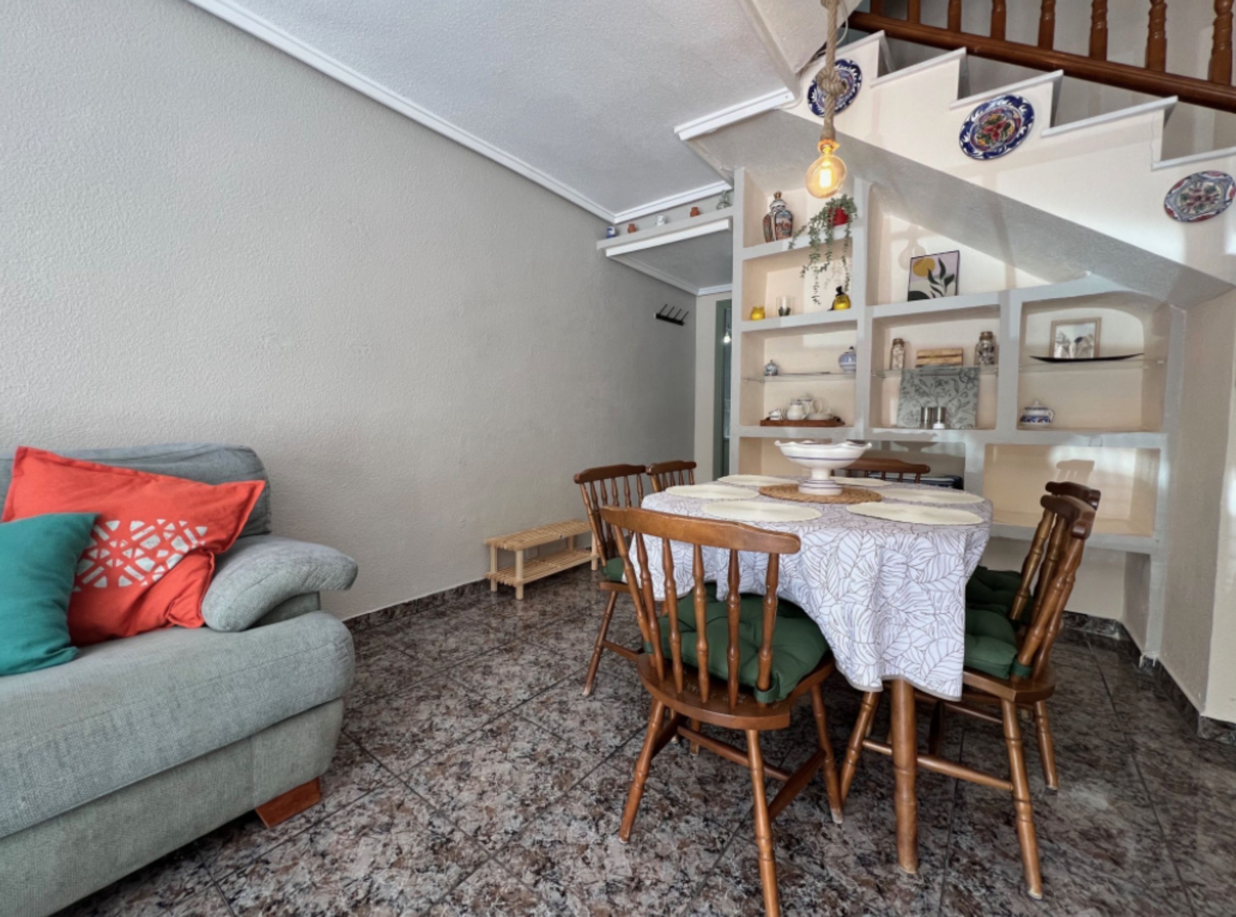 A dining area with a table set for four, surrounded by wooden chairs with green cushions. A grey sofa with red and teal cushions is against the wall. The room has a tiled floor and a sloped ceiling with a hanging light fixture.