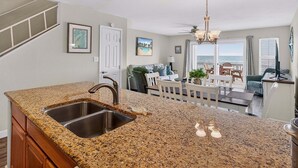 A kitchen with granite countertops, a stainless steel sink, and a ceiling fan. There is a dining area with a table and chairs, and a view of the ocean through large windows. - Kontiki Bayfront K304 3 Bedroom Condo (Rockport)