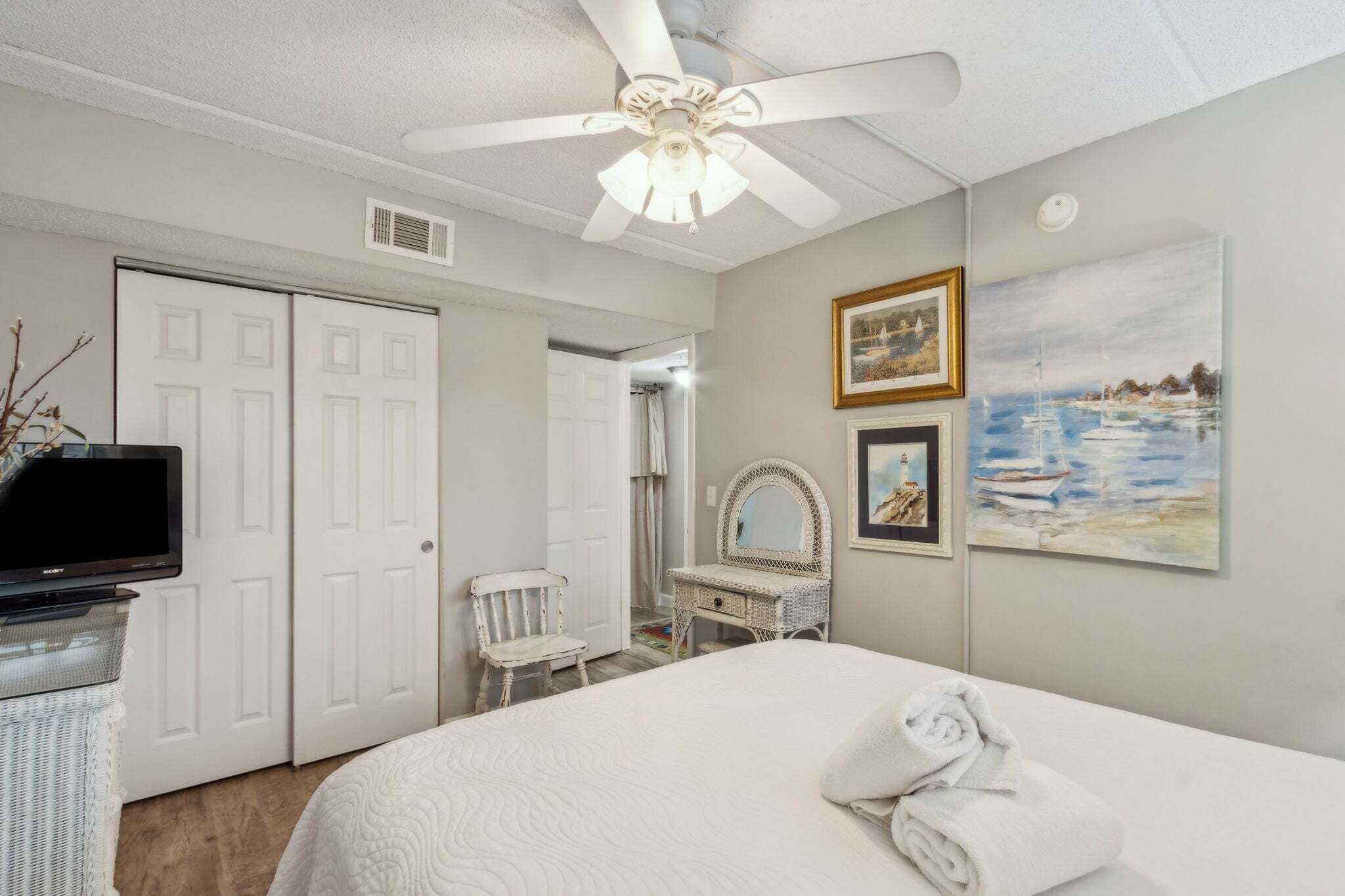 A bedroom with a bed, a ceiling fan, a television, a chair, a vanity with a mirror, and artwork on the wall.