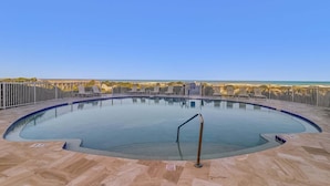 Une grande piscine extérieure avec une rambarde en métal, un poste de surveillance pour les sauveteurs et un ciel d’un bleu limpide.