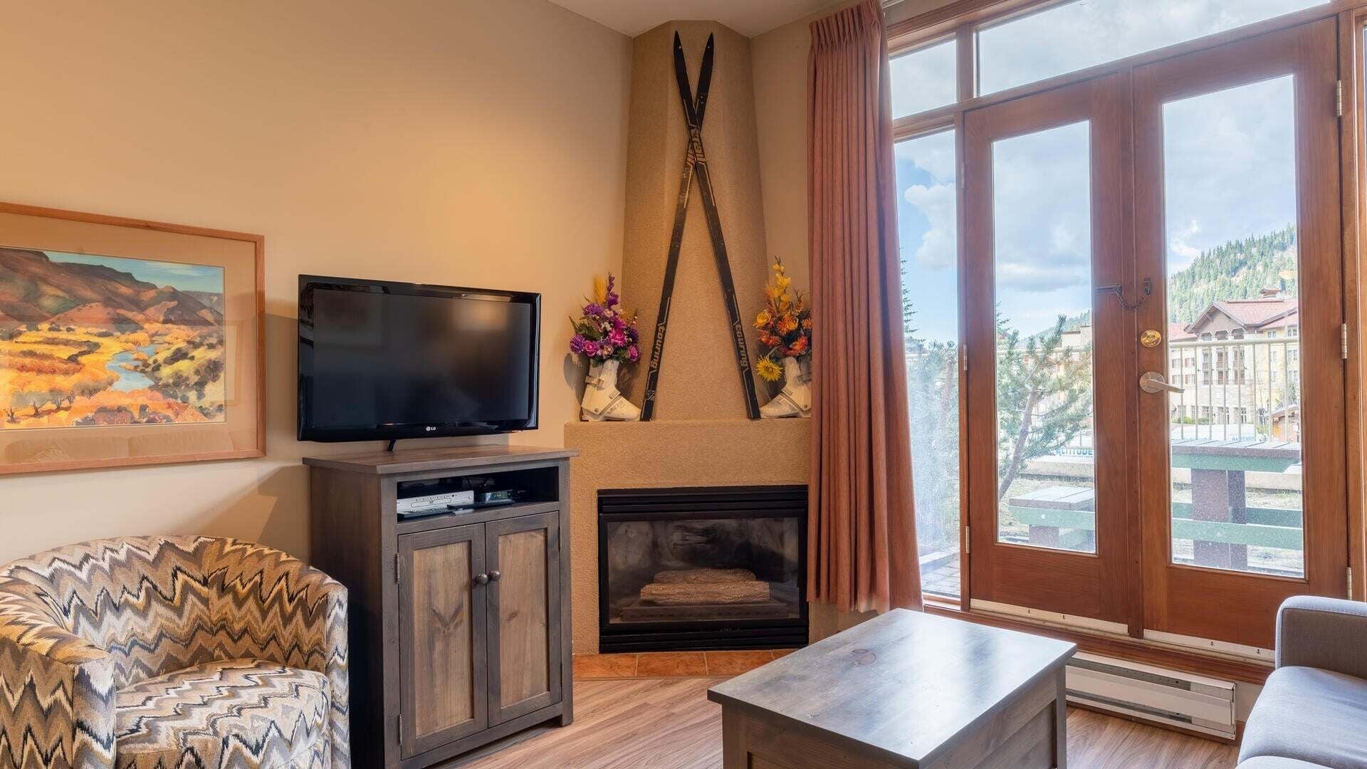 A living room with a television, a fireplace, and a large window with a view of a building and trees.