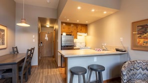 A compact kitchen with wooden cabinets, a stainless steel refrigerator, a stove, and a sink. There is a dining table with chairs and a bar stool.