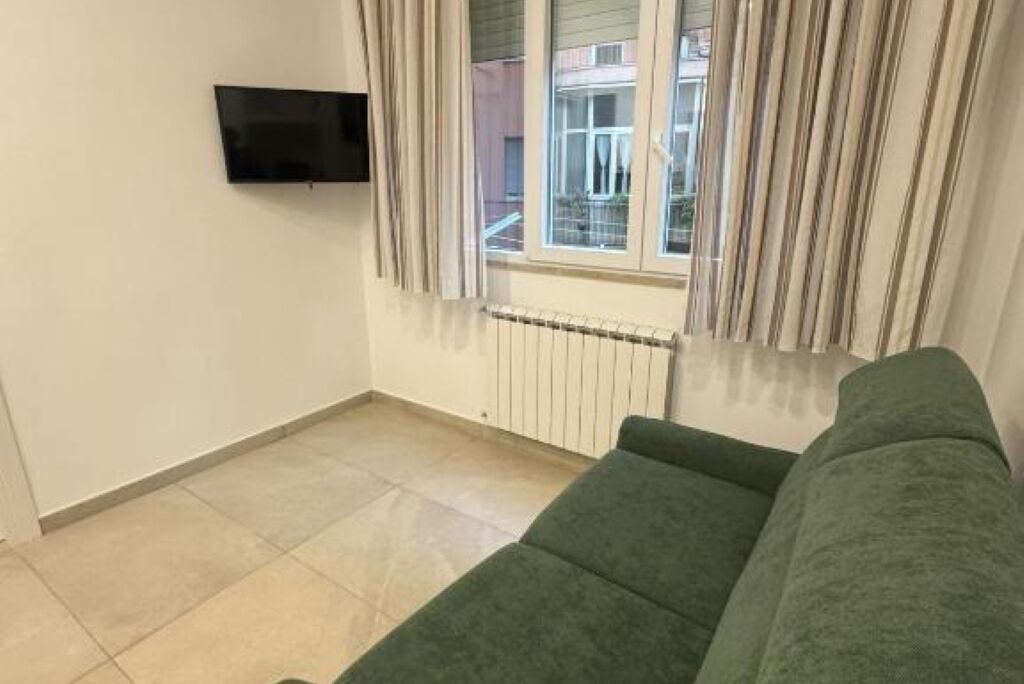 Ett vardagsrum med en grön soffa, en platt-TV som är monterad på väggen och ett fönster med gardiner.