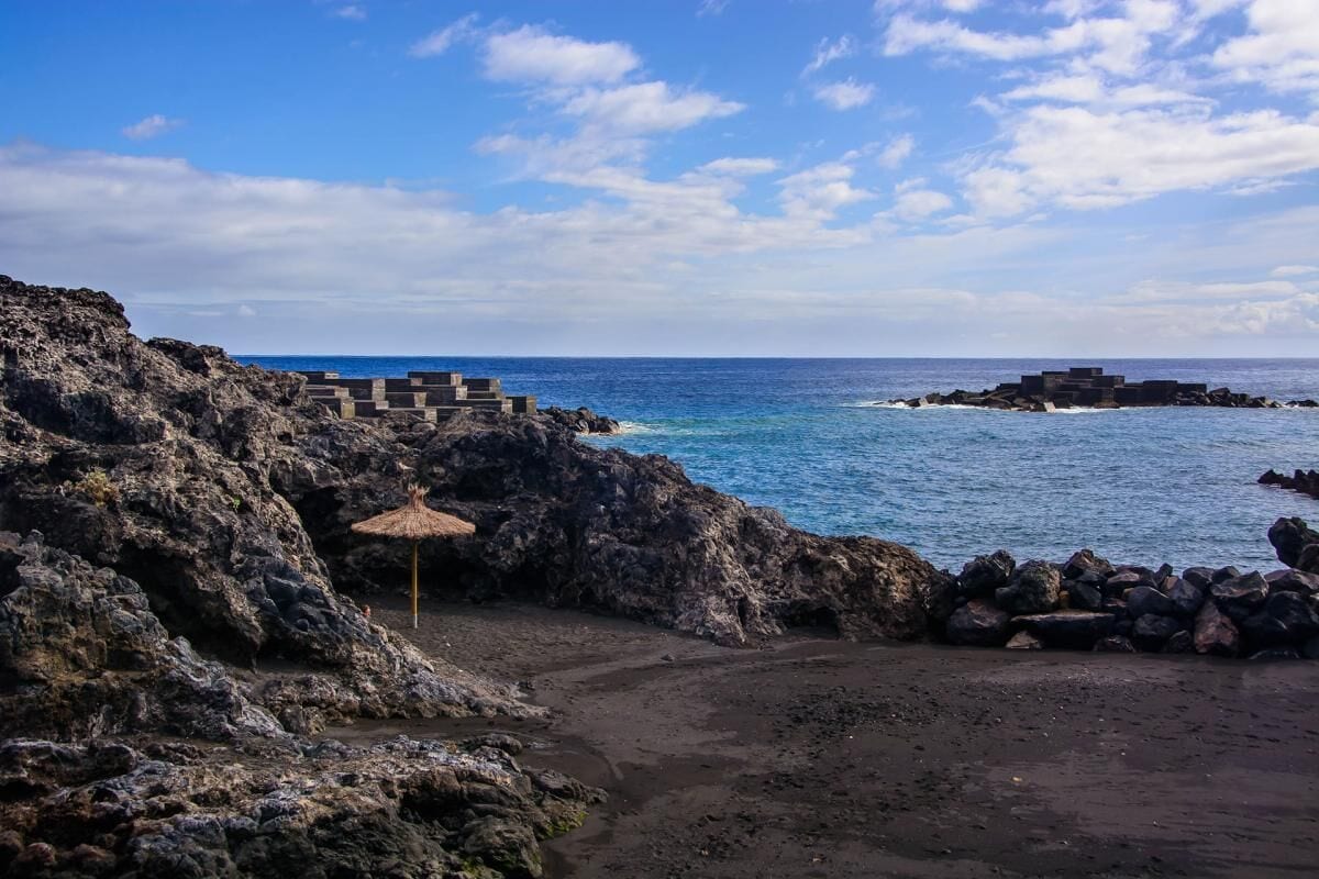 Paisagem costeira rochosa com céu claro, uma praia e uma construção feita de blocos de pedra.
