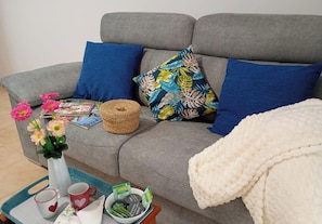 A grey sofa with blue and floral cushions, a woven basket, a vase with pink flowers, a tray with cups and tea bags, and a white blanket. - Charming 1-bedroom Apartment With Sea Views in Breña Baja (Brena Baja)