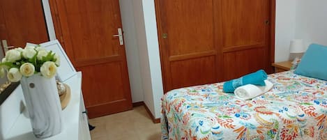 Un dormitorio con una cama cubierta por una colcha floral colorida, un tocador blanco con un jarrón de flores y puertas dobles de madera.