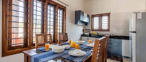 Un coin repas avec une table en bois dressée pour le petit-déjeuner, une cuisine équipée d’appareils électroménagers modernes et de grandes fenêtres à cadres en bois.