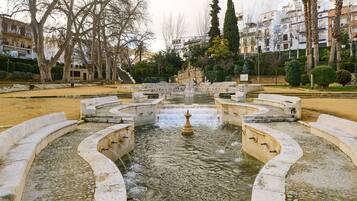 A fountain with a central statue, surrounded by stone steps and bordered by trees and buildings.