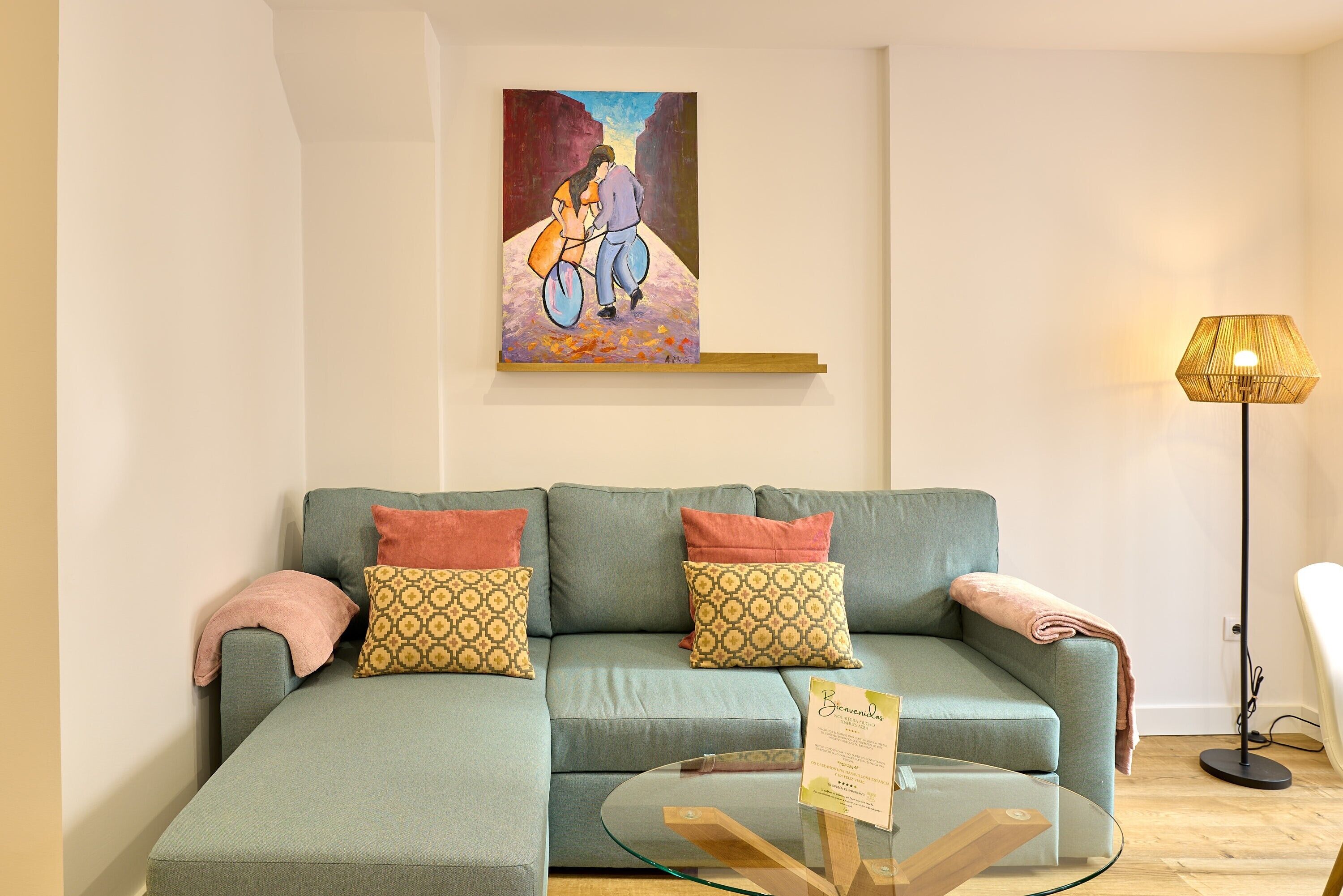 Una sala moderna con un sofá modular, cojines estampados, una mesa de centro de vidrio, un cuadro en la pared y una lámpara de pie.