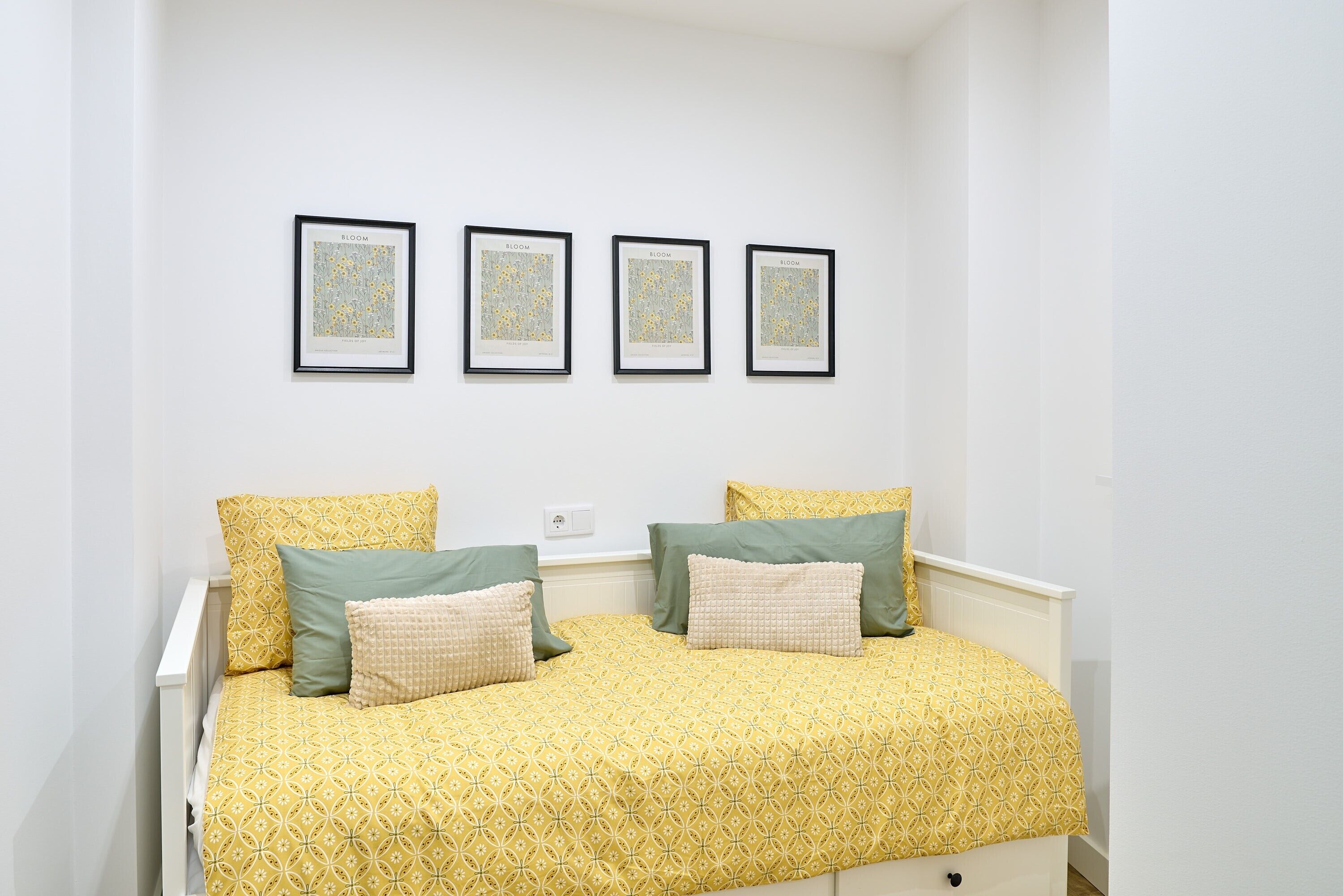 A bed with a yellow patterned bedspread, two pillows, and a white headboard. There are three framed pictures on the wall above the bed.