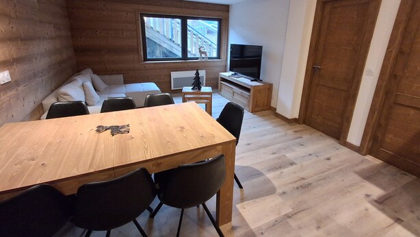 Una sala de estar moderna con una mesa de comedor de madera, sillas negras, un sofá, un televisor y una ventana con vistas a un edificio.