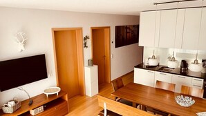 A modern kitchen with wooden cabinets, a dining area with a wooden table and benches, a TV on a wooden stand, and a white wall with a decorative item. - House Annabelle Top 1 (Damüls)
