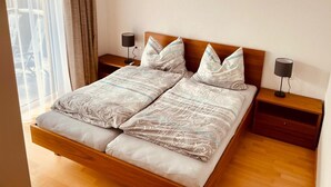 A double bed with patterned bedding, two bedside tables with lamps, a hanging light fixture, and a view of a balcony through sheer curtains. - House Annabelle Top 1 (Damüls)