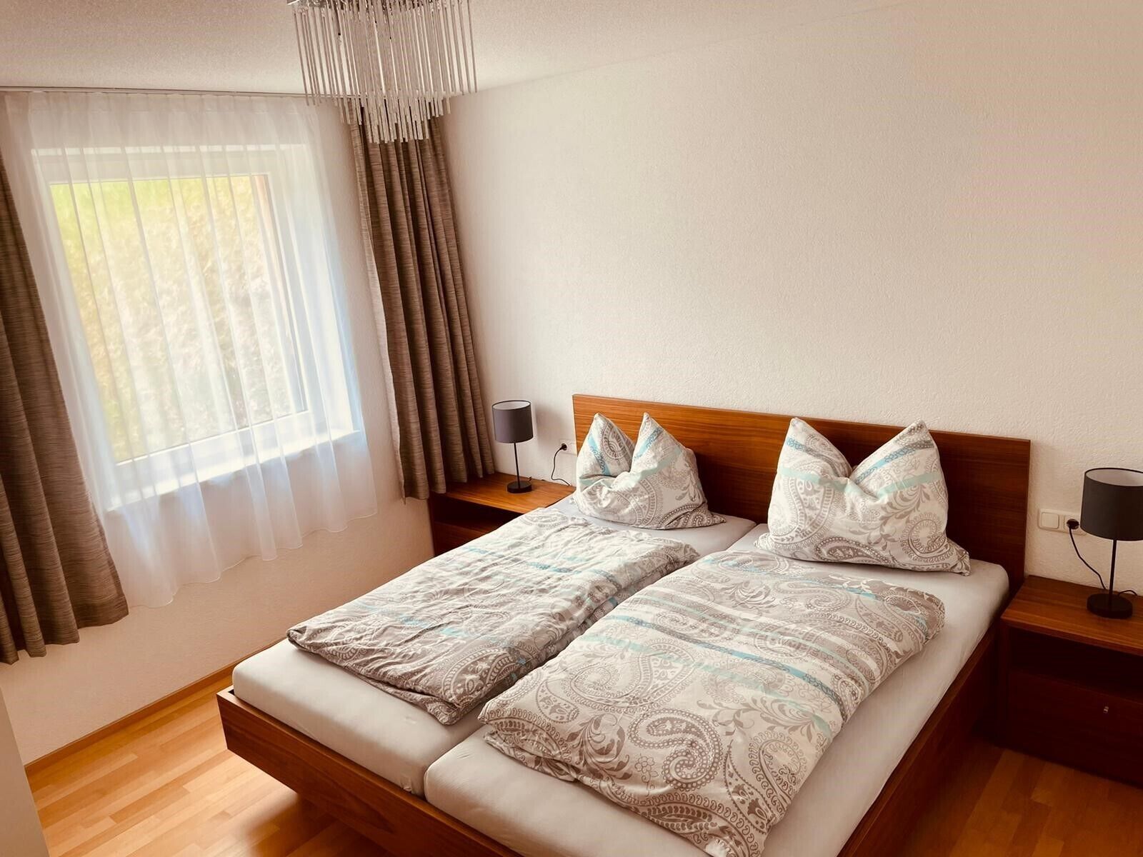 A double bed with patterned bedding, a wooden headboard, bedside tables with lamps, and a window with sheer curtains and a draped valance.