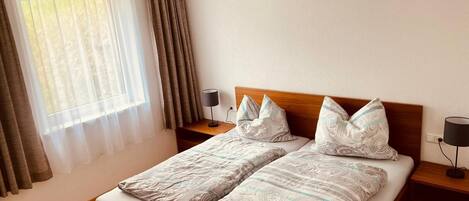 A double bed with patterned bedding, a wooden headboard, bedside tables with lamps, and a window with sheer curtains and a draped valance.