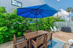 An outdoor patio with a wooden table and chairs, a blue umbrella, and a white fence in the background. - Cute Port Aransas home near the beach, shops, and restaurants. Community pool an (Port Aransas)