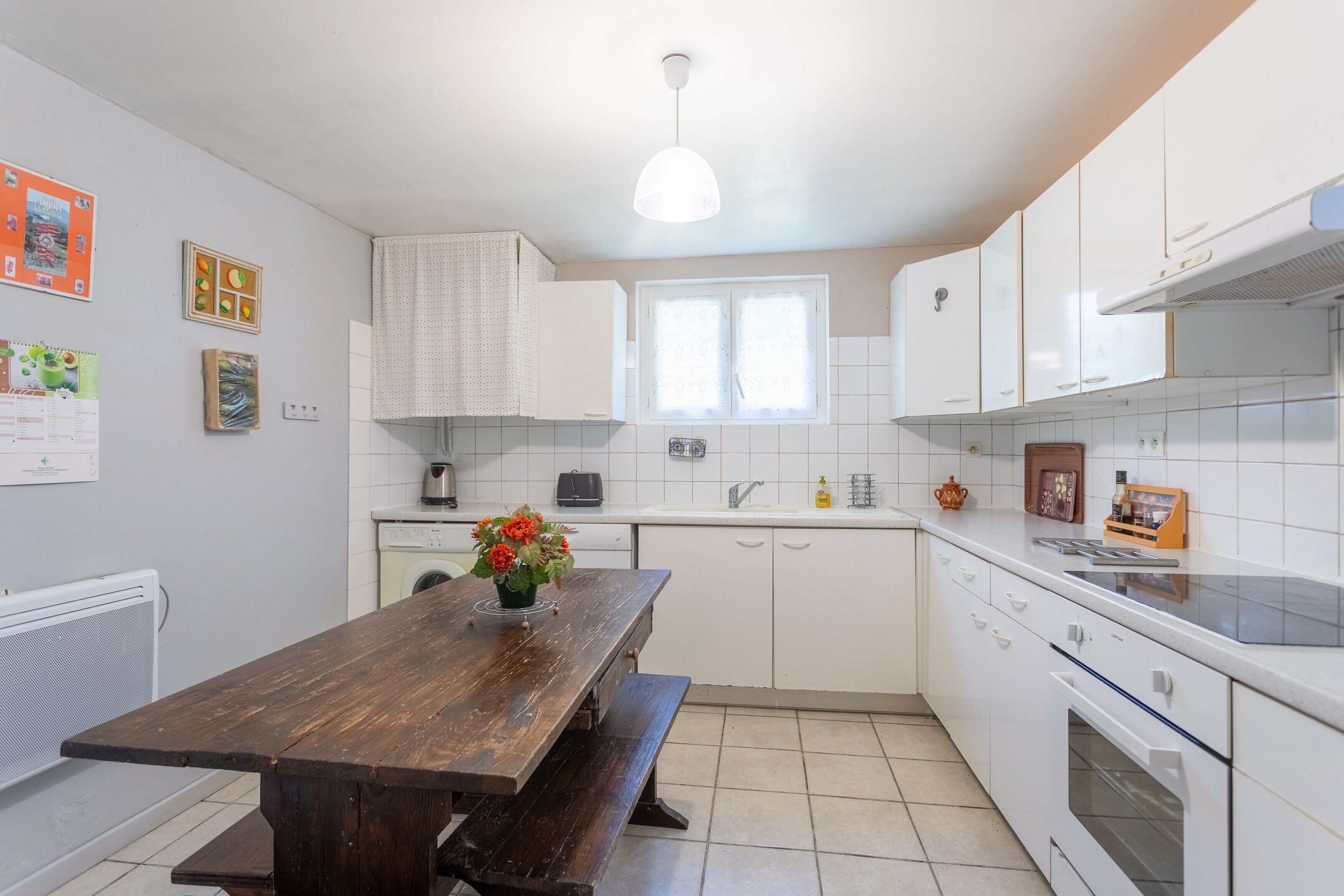 Une cuisine avec des meubles blancs, une table en bois accompagnée d’un banc, une fenêtre et un luminaire suspendu.