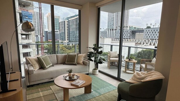 A modern living room with a sofa, armchair, and coffee table, large windows with a city view, and a small balcony with a chair and table. - Sophisticated 2BR/2BA at Miami World Center (Miami)