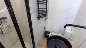 Un baño moderno con cabina de ducha de vidrio, toallero y cubo de basura negros, y un dispensador de jabón sobre una encimera de madera.