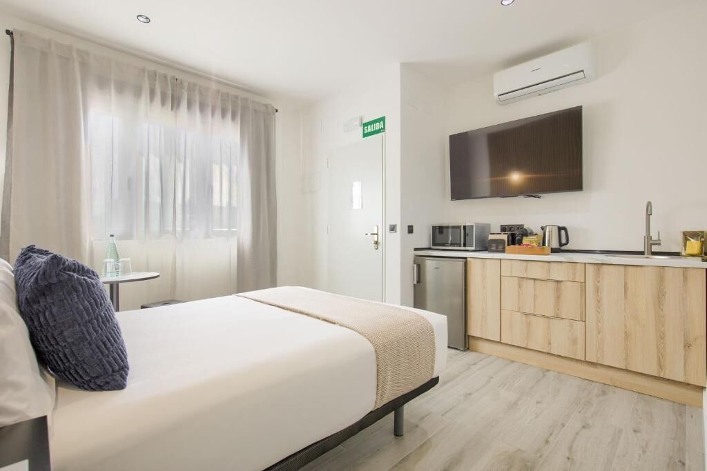 A hotel room with a bed, a small kitchenette, a TV mounted on the wall, and a sign indicating the exit.