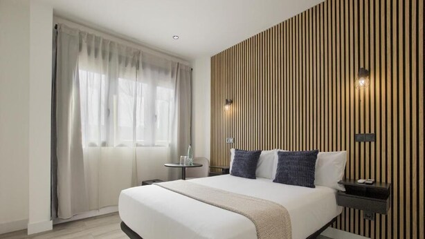 Una habitación de hotel moderna con una cama grande, una mesita de noche, una pequeña mesa y una ventana con cortinas.