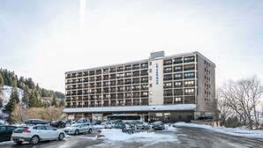 A multi-story hotel with a central entrance, parking lot, and a view of a snowy landscape. - Cute studio apartment for 4 people, balcony and garage (Chamrousse)