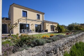 A two-story house with a stone wall in front, a patio with outdoor furniture, and a garden with various plants.