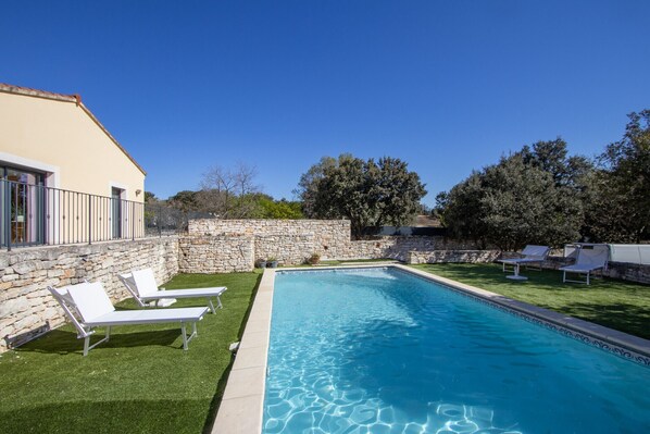 A swimming pool with lounge chairs, a stone wall, and a building with a sloped roof.