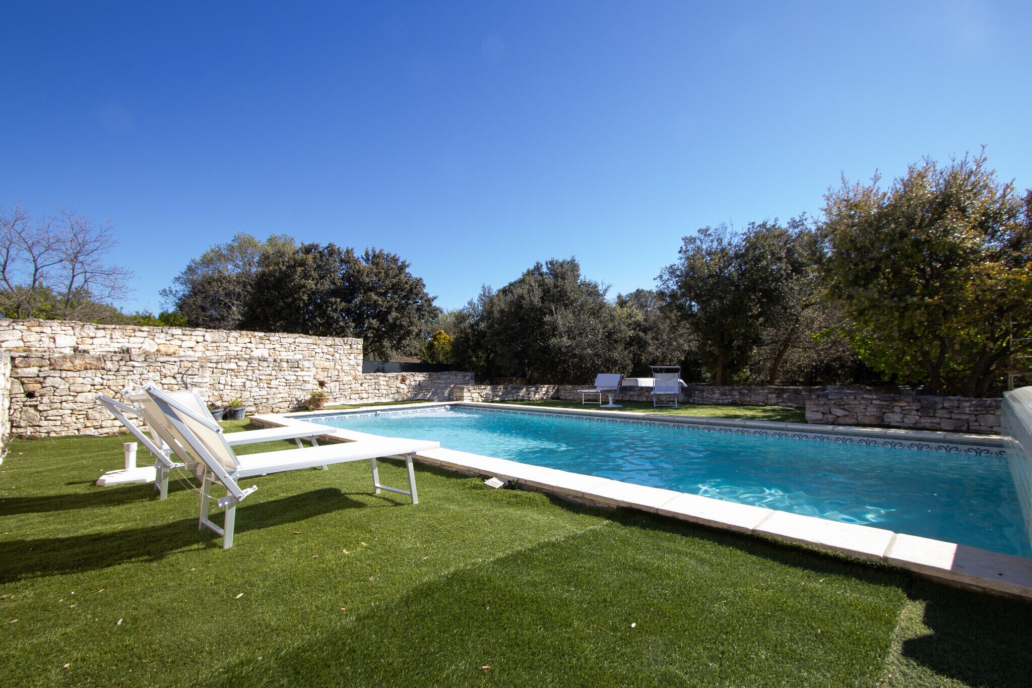 Une piscine avec des chaises longues, un mur en pierre et de l’herbe verte.