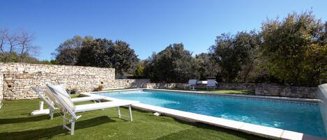 Une piscine avec des chaises longues, un mur en pierre et de l’herbe verte.