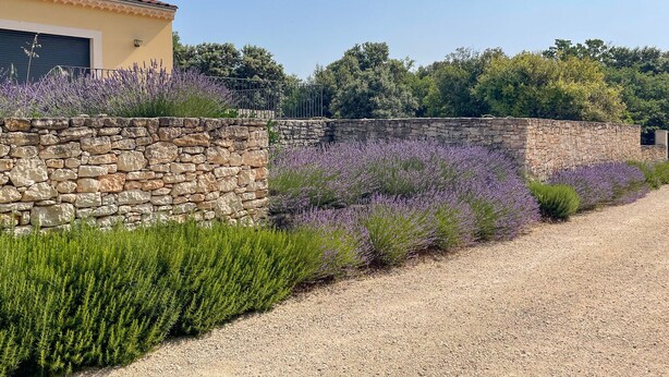 A stone wall with lavender plants and a gravel path leading to a building with a closed garage door. - Air-conditioned villa with private pool – Wheelchair accessible – Drôme Provençale (Saint-Restitut)