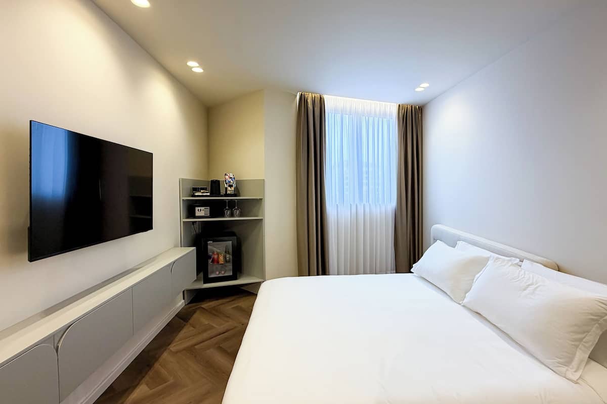 A hotel room with a flat-screen TV, a minibar, a bed, and a window with curtains.