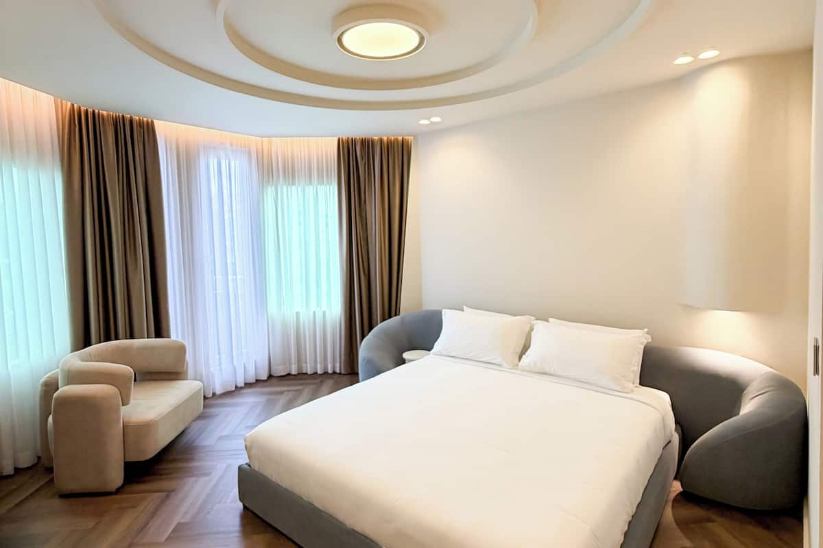 A modern bedroom with a large bed, two armchairs, and a curved ceiling with recessed lighting.