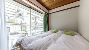 Une chambre avec une grande fenêtre, un lit recouvert d’une literie blanche et deux oreillers verts.
