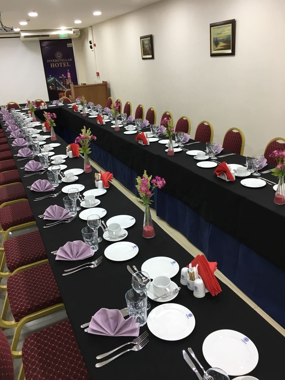 Una sala conferenze con un lungo tavolo apparecchiato per una riunione, completa di sedie, piatti, posate e centrotavola floreali.