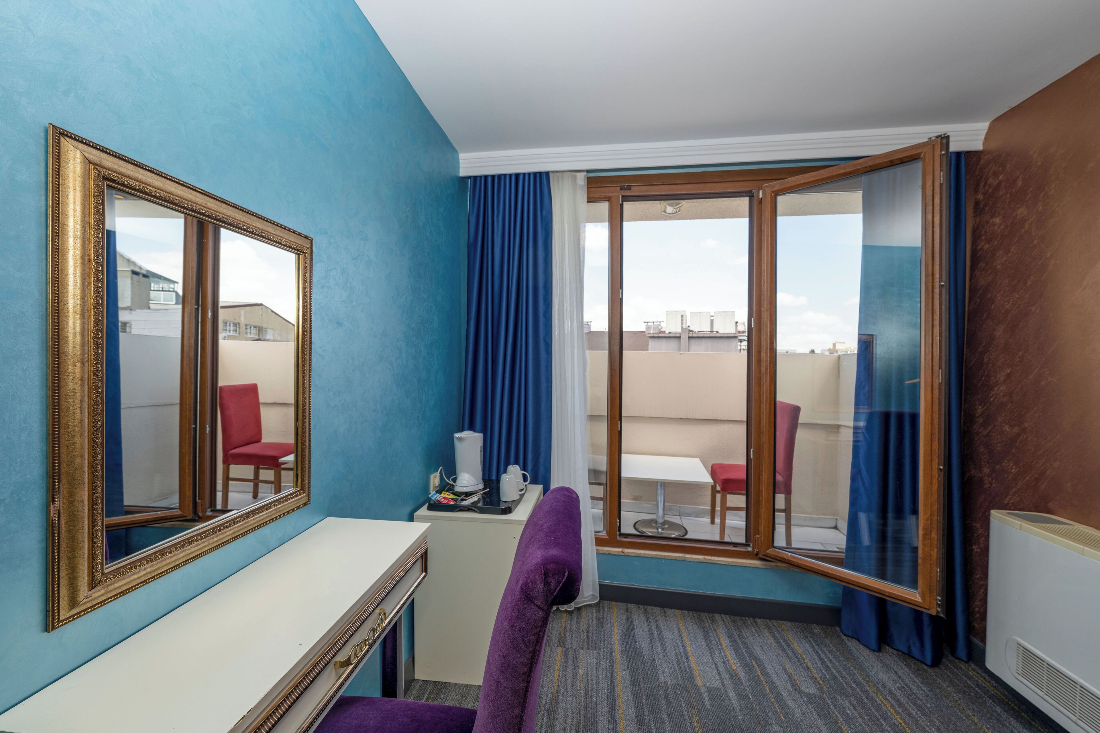 A hotel room with a large mirror, a desk, a purple chair, a small table with a white mug, and a view through a sliding glass door to an outdoor seating area.