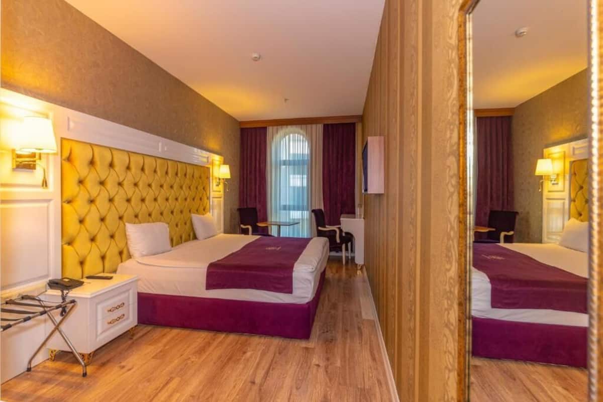 A hotel room with a tufted headboard, two beds with purple bedspreads, a bedside table with a lamp, a desk with a chair, and a large mirror.